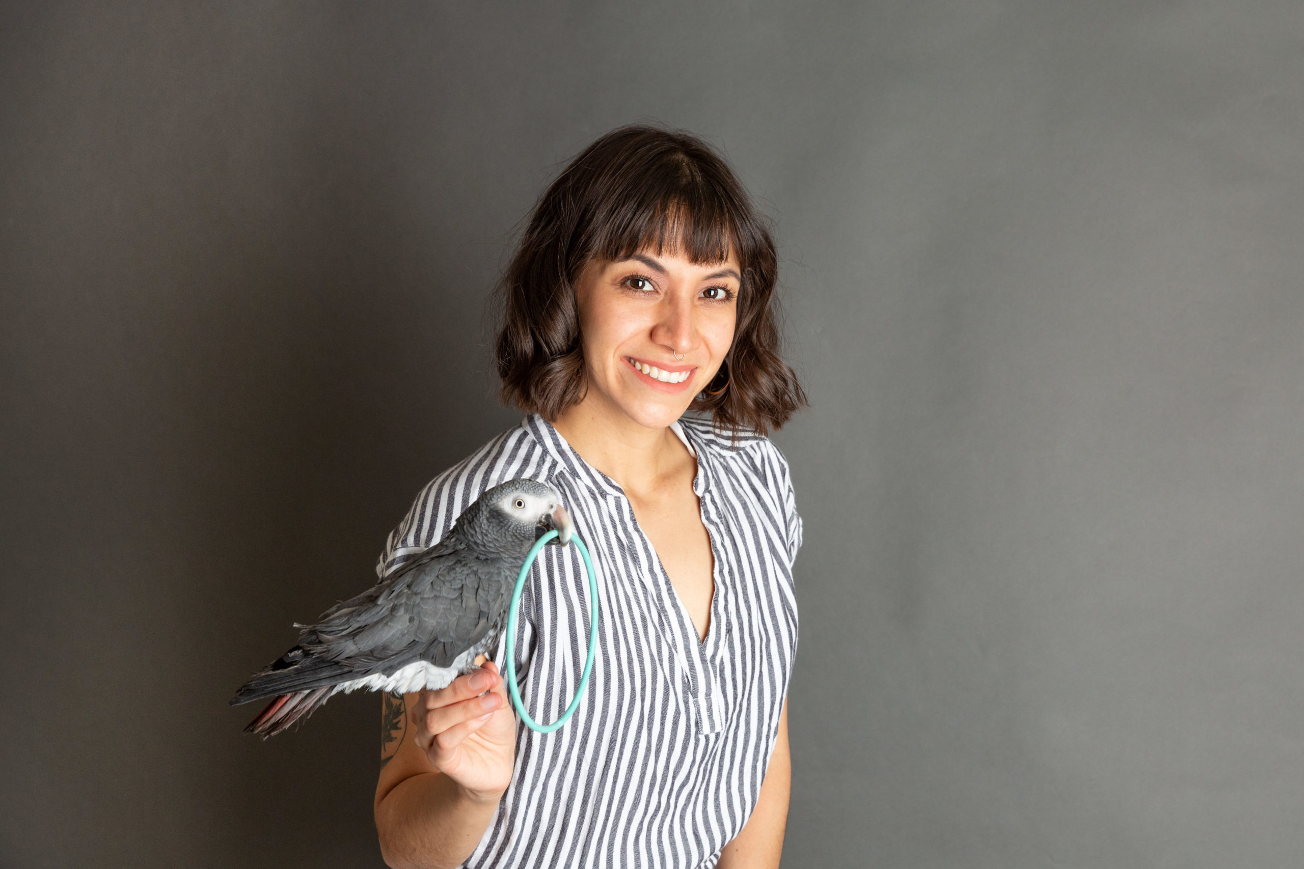 Amelia, with shoulder-length brown hair and a striped shirt smiles with a parrot posed on her hand.