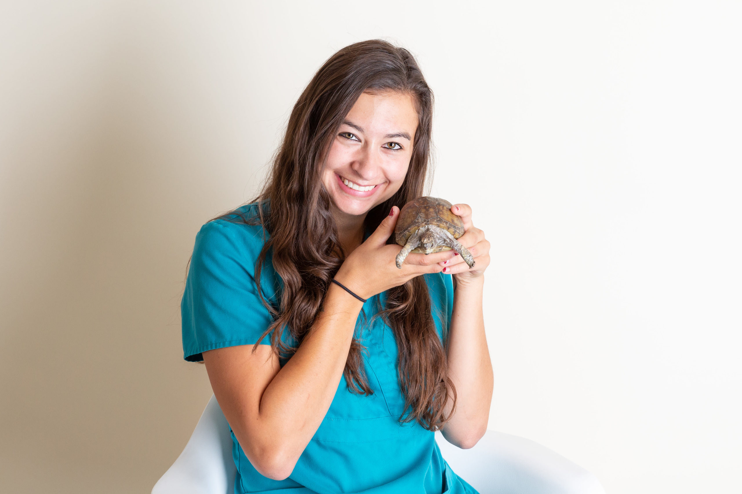 Bre, with long dark hair, wearing teal scrubs, is holding up a turtle and smiling.